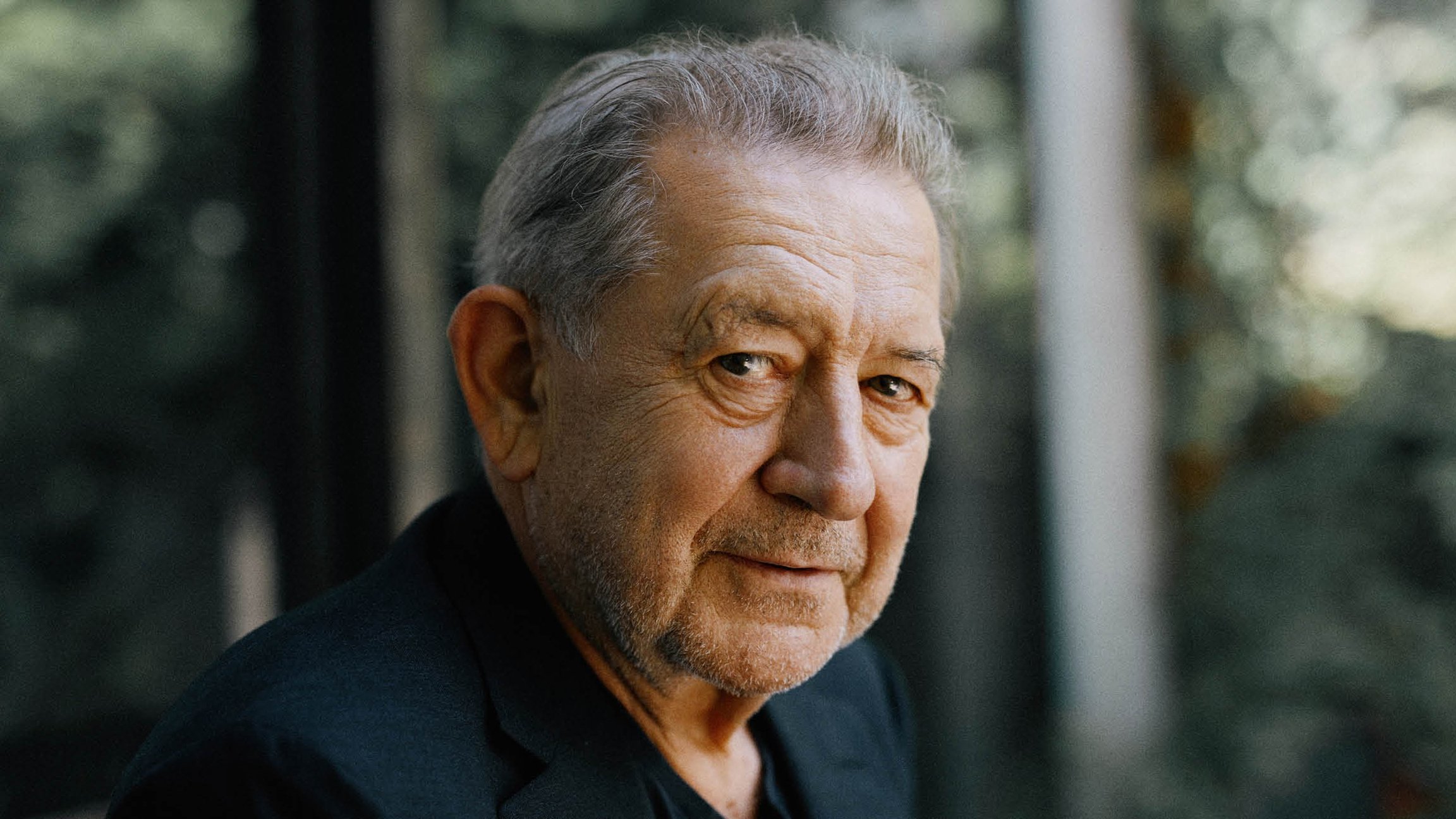 Portrait von André Jung in einem schwarzen Sakko seitlich in die Kamera blickend, der Hintergrund verschwommen