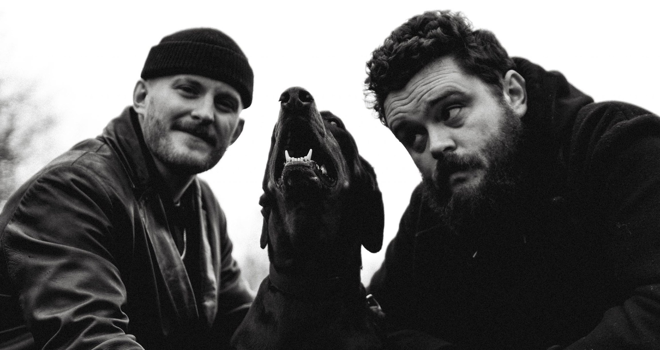 Schwarz-weiss Bild mit zwei dunkel gekleideten Männern und einem Hund