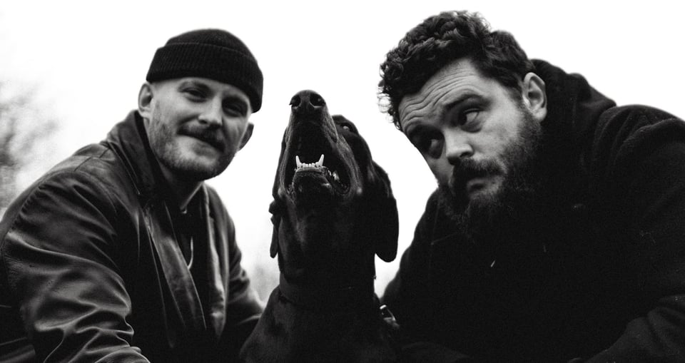 Schwarz-weiss Bild mit zwei dunkel gekleideten Männern und einem Hund