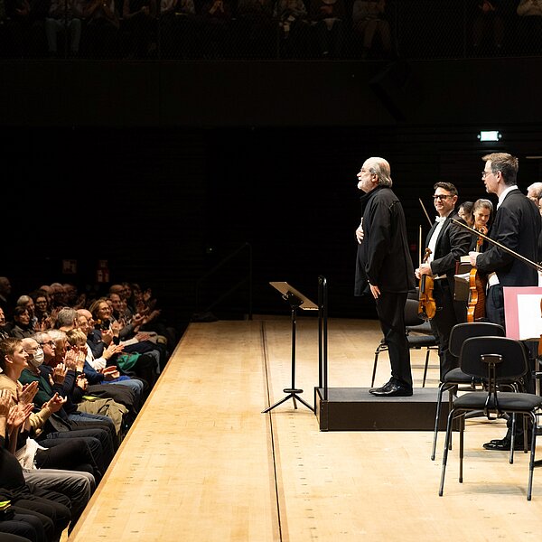 Ein Dirigent steht vor seinem Orchester auf einer Bühne vor einem klatschenden Publikum, er ist seitlich zu sehen. 