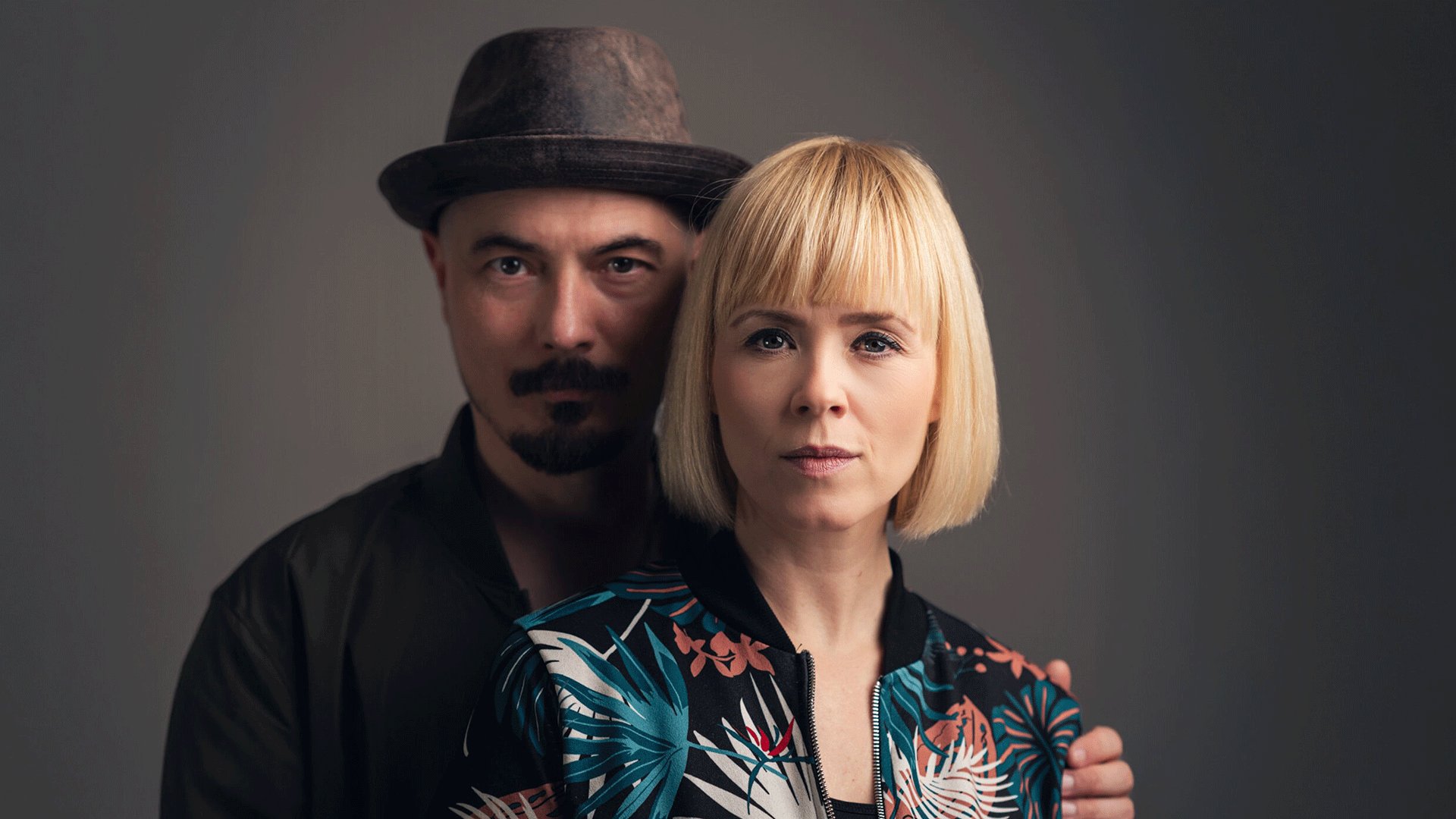 Ein Portrait vom Duo Carolin No vor einem grauen Hintergrund eine Person mit Hut steht hinter einer Person mit blondem Bob und gemusterter Jacke.