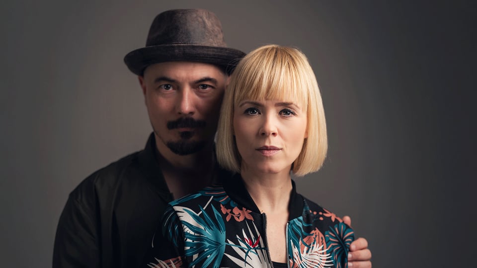 Ein Portrait vom Duo Carolin No vor einem grauen Hintergrund eine Person mit Hut steht hinter einer Person mit blondem Bob und gemusterter Jacke.