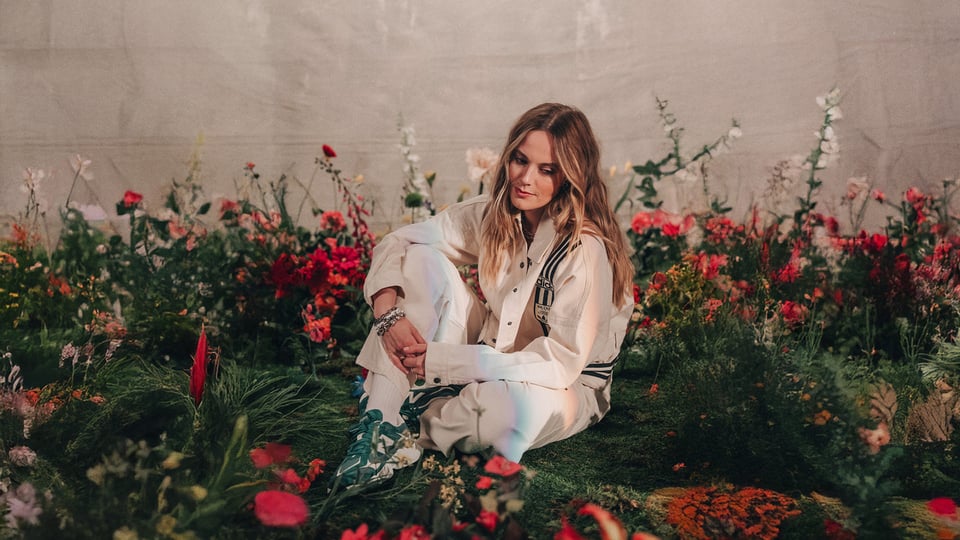 Foto von der Künstlerin Sophia, sitzend in einem Garten mit Blumen.