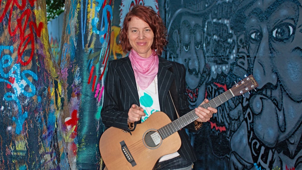 Eine Person mit dunkler, gestreifter Jacke und Akustikgitarre steht vor bunt besprühten Graffiti‑Wänden; die Gitarre wird mit beiden Händen gehalten, während farbenfrohe Muster und Figuren die Hintergrundflächen bedecken