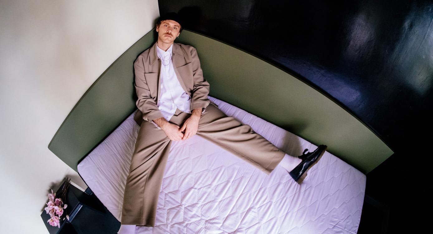 Der Künstler in einem braunen Anzug und mit Mütze, sitzend in der Ecke auf einem Bett.