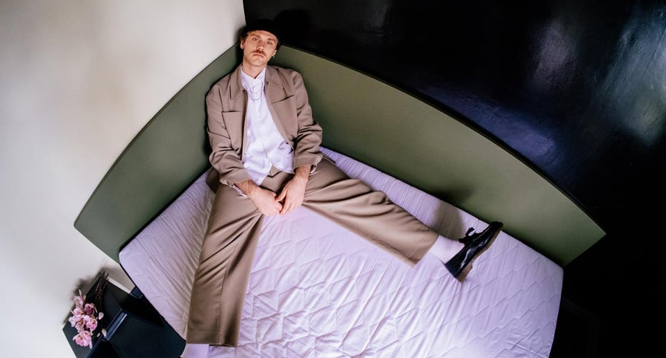Der Künstler in einem braunen Anzug und mit Mütze, sitzend in der Ecke auf einem Bett.