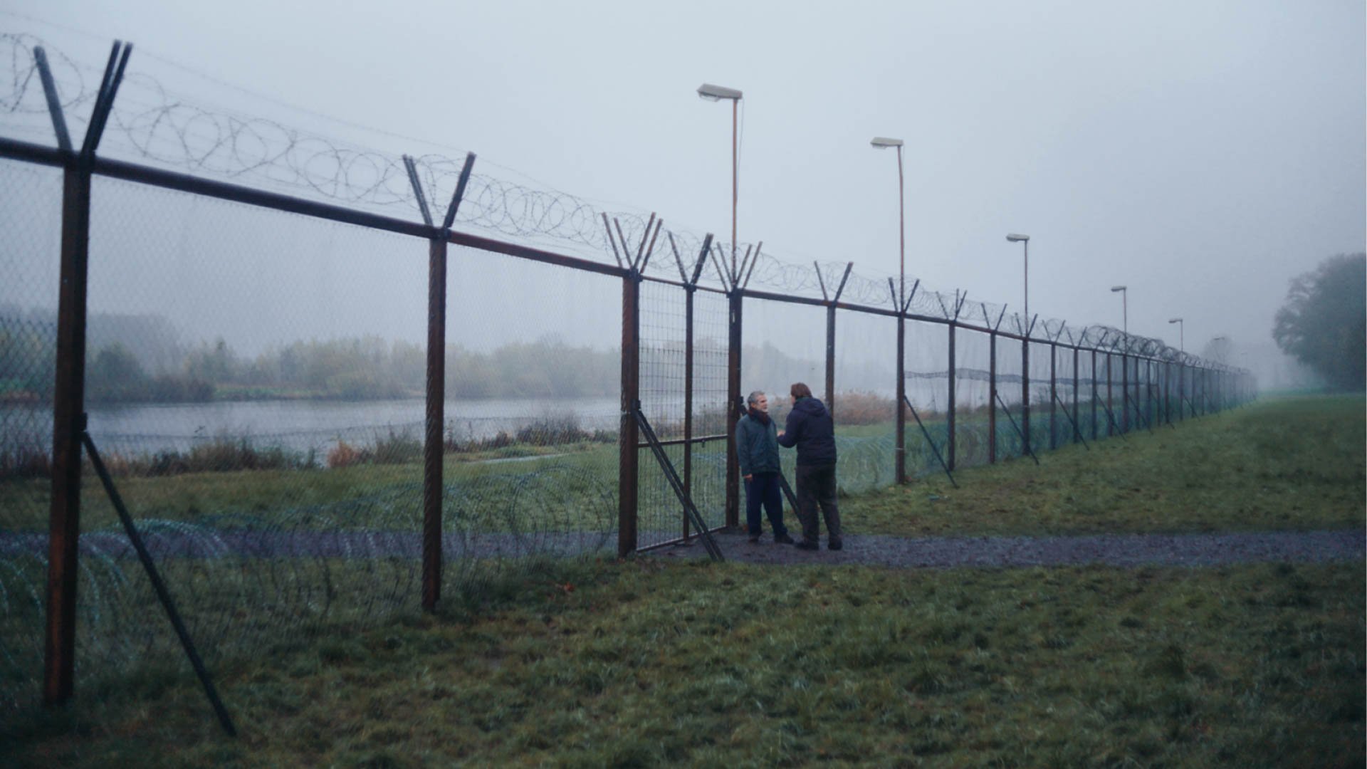 Zwei Personen stehen in einer nebligen Landschaft vor einem langen Zaun mit Stacheldraht, daneben verläuft ein Gewässer.