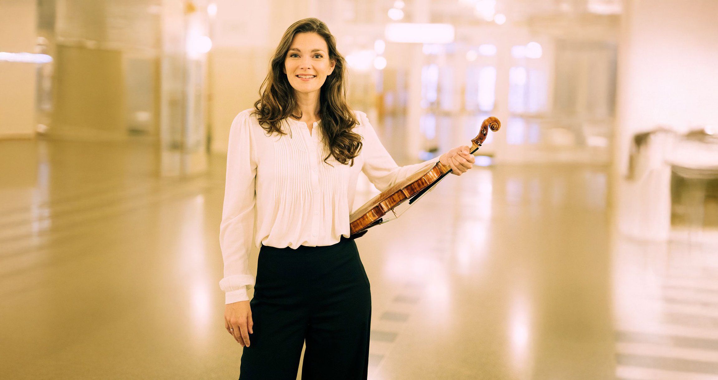 Dame mit Violine in weisser Bluse und schwarzer Hose stehend vor hellem Hintergrund