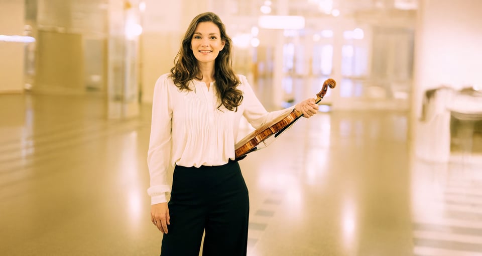 Dame mit Violine in weisser Bluse und schwarzer Hose stehend vor hellem Hintergrund