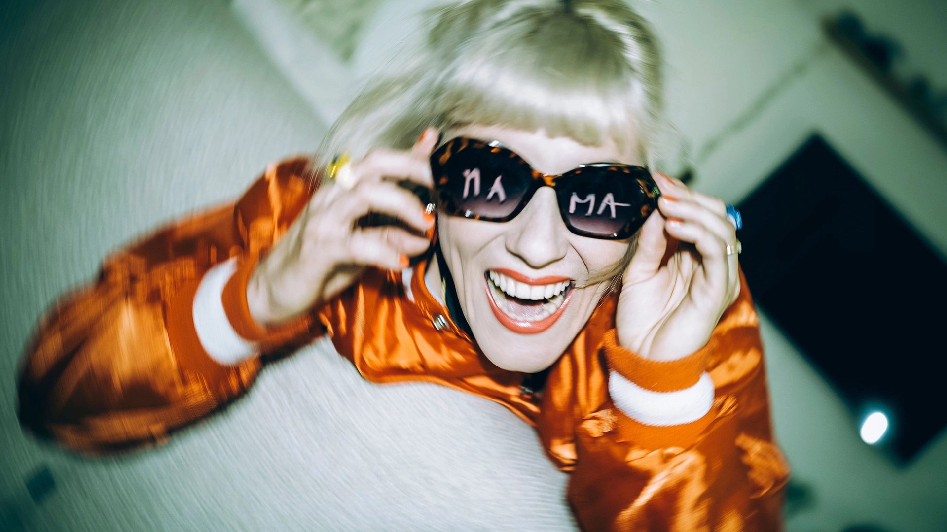 Person mit blondem Haar und Sonnenbrille lächelt in die Kamera und trägt eine glänzende orange Jacke; in den Brillengläsern ist Schrift zu sehen, Aufnahme mit unscharfem Hintergrund und starker Bewegung.