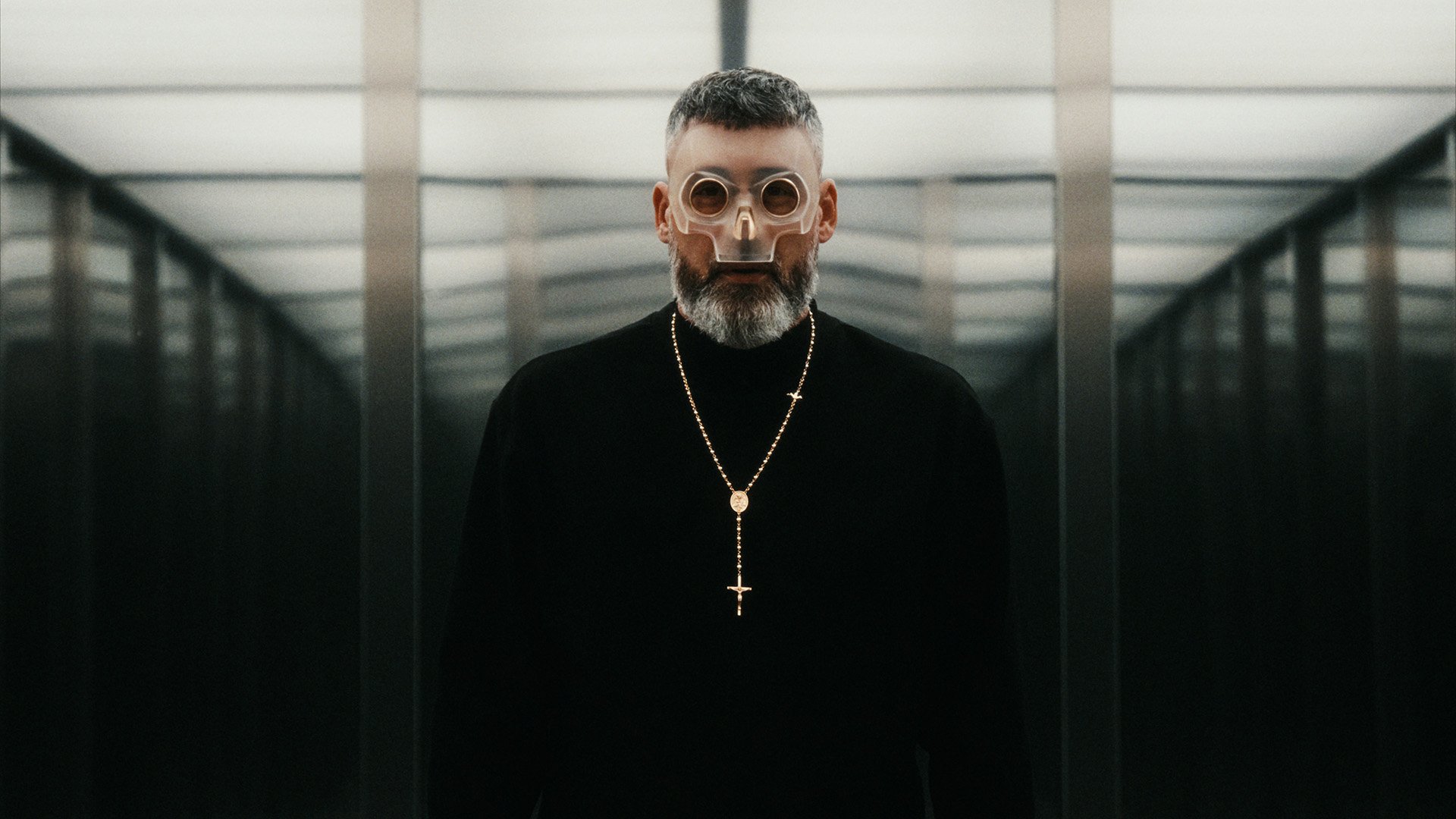 Sido mit transparenter Maske und Kreuzkette steht in einem symmetrischen, spiegelnden Gang mit futuristischer Atmosphäre.