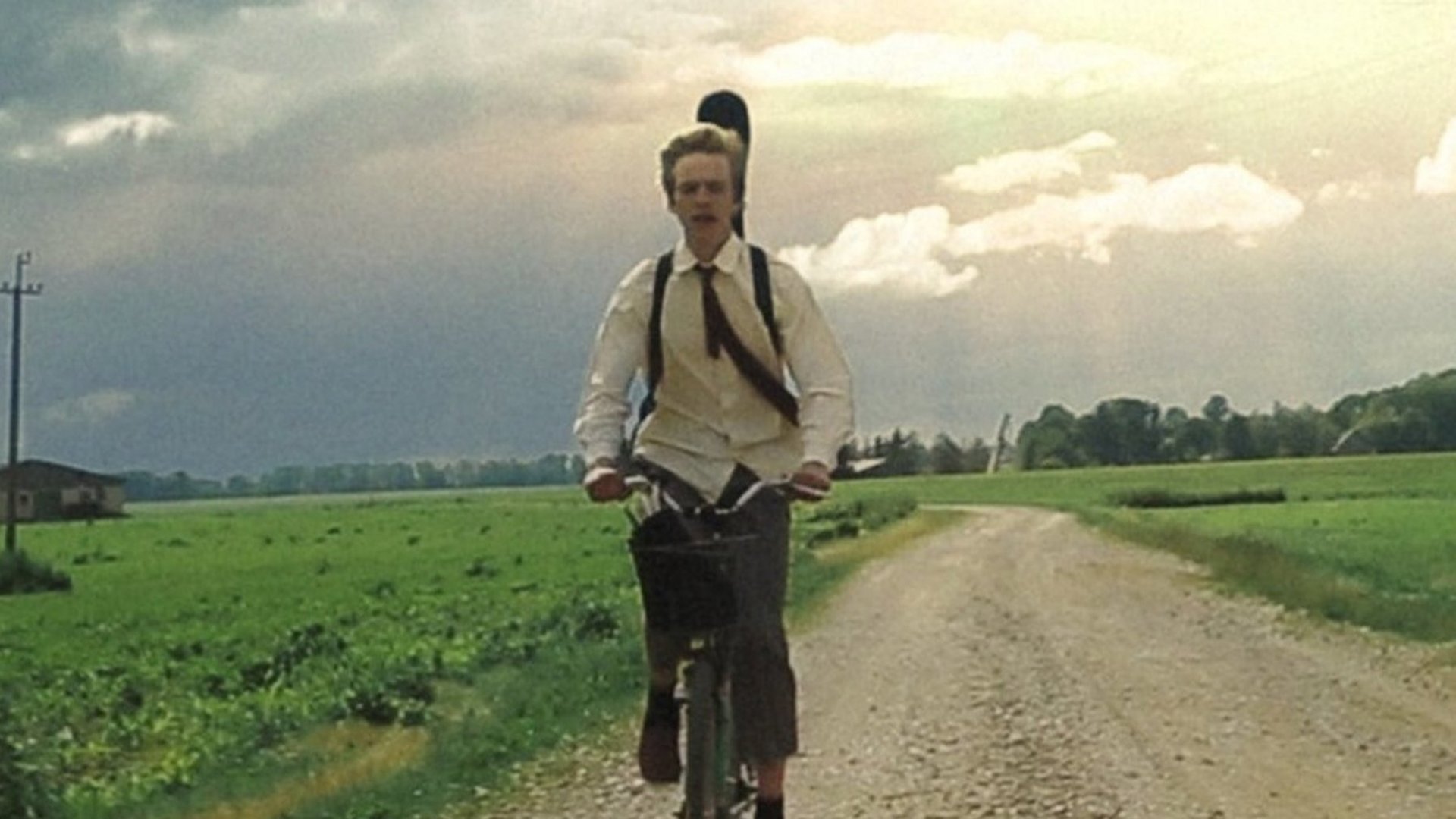 Ein junger Mann auf einem Feldweg auf dem Fahrrad mit einem Instrumentenkoffer auf dem Rücken