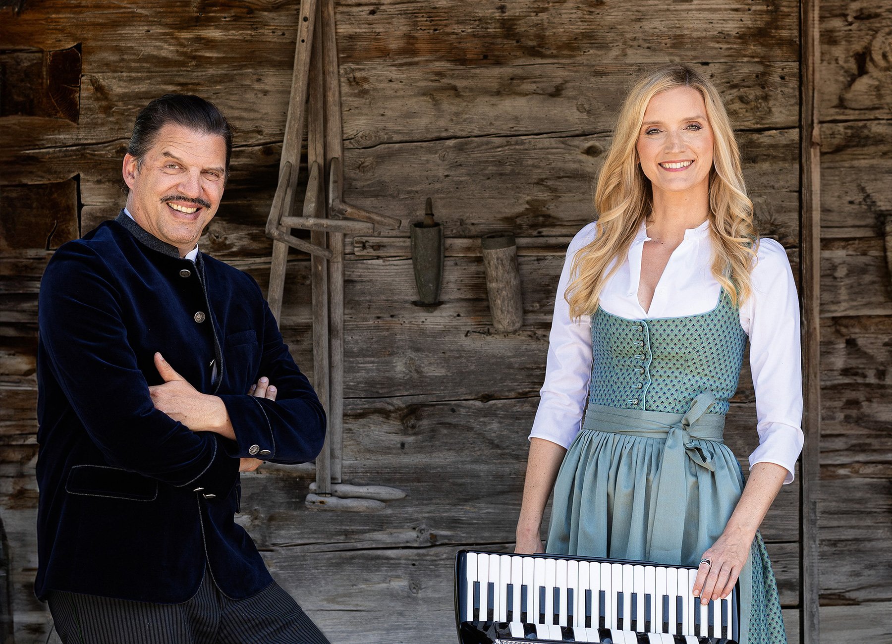 Eine Dame mit Instrument und ein Herr, beide in Tracht vor hölzernem Hintergrund