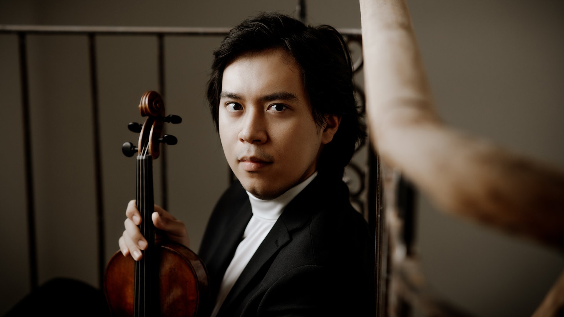 Porträt des Violinisten Elias David Moncado in schwarzer Konzertkleidung mit Violine auf einer Treppe. Der Musiker blickt direkt in die Kamera, während warmes Licht eine ruhige, klassische Atmosphäre erzeugt.