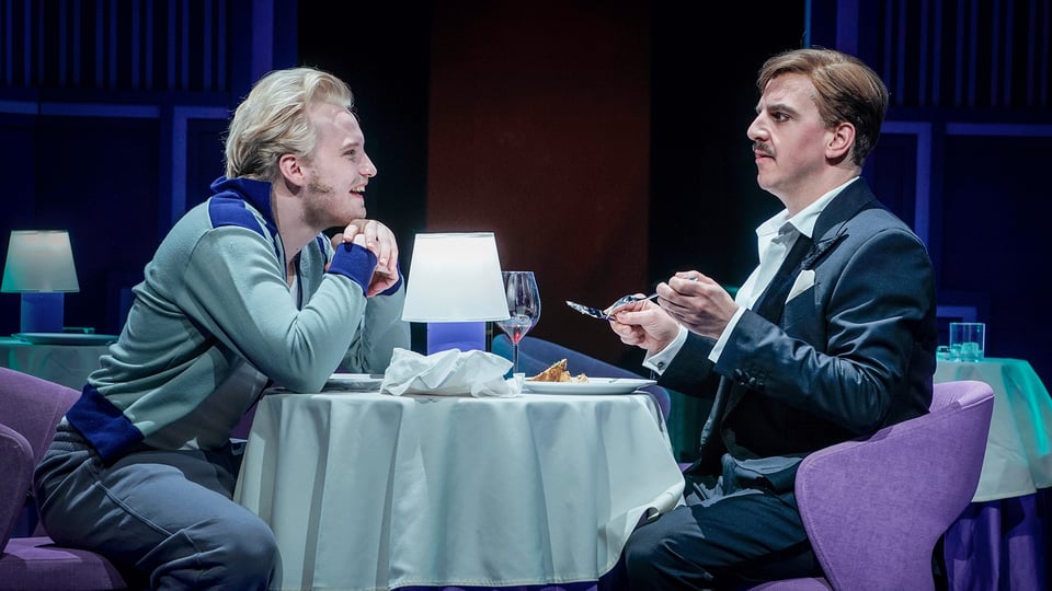 Theaterszene in einem Restaurant. Zwei Darstellende sitzen sich an einem gedeckten Tisch mit Tischlampe, Weinglas und Dessert gegenüber, während eine Person im Anzug mit Besteck in der Hand spricht und die andere sich über den Tisch lehnt.