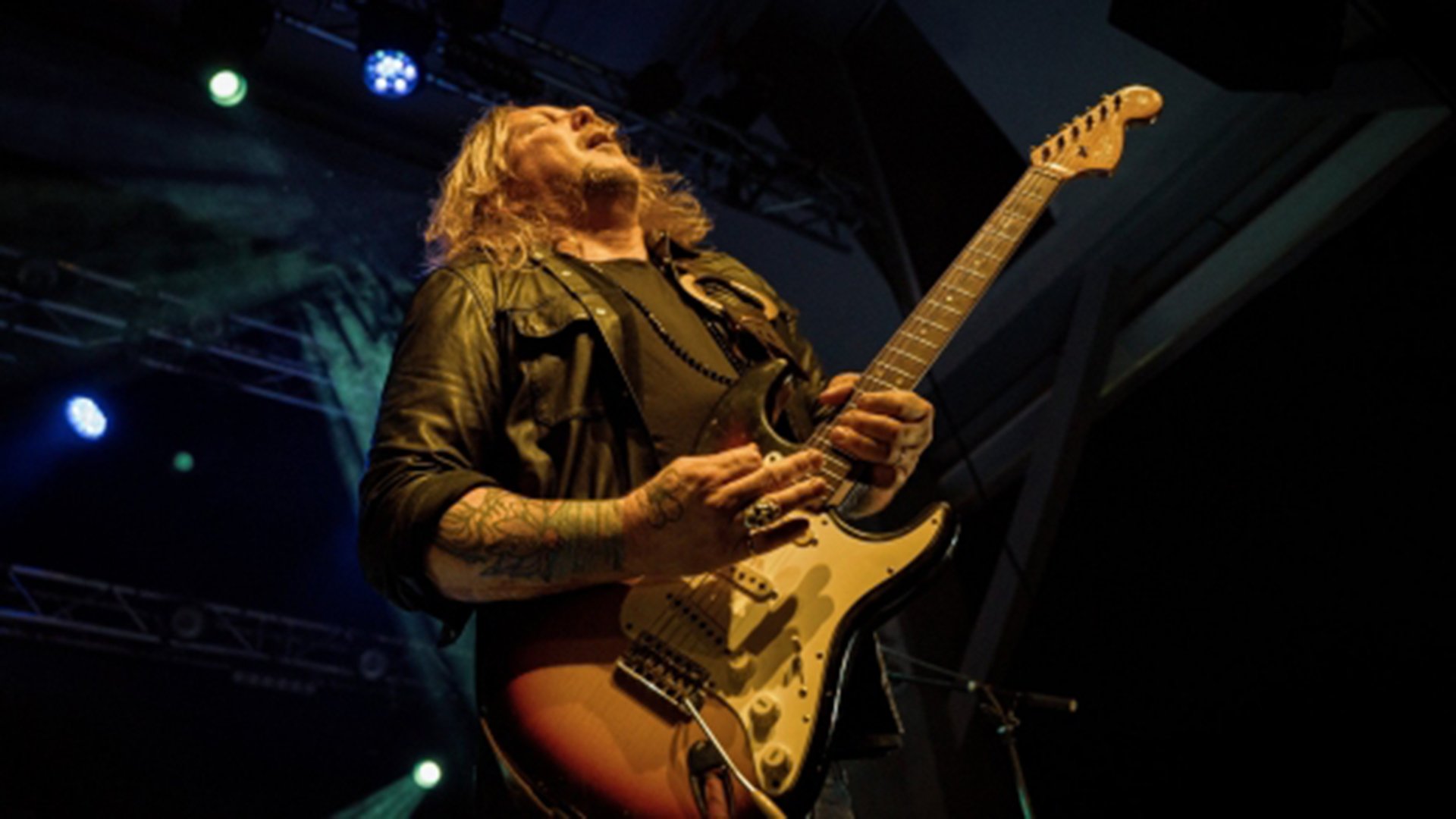 Ein Künstler mit Gitarre auf der Bühne, in warmen orangem Licht, von oben Scheinwerfer, die herableuchten