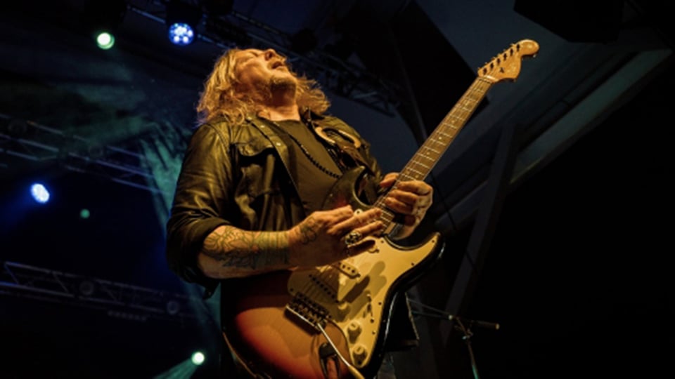 Ein Künstler mit Gitarre auf der Bühne, in warmen orangem Licht, von oben Scheinwerfer, die herableuchten