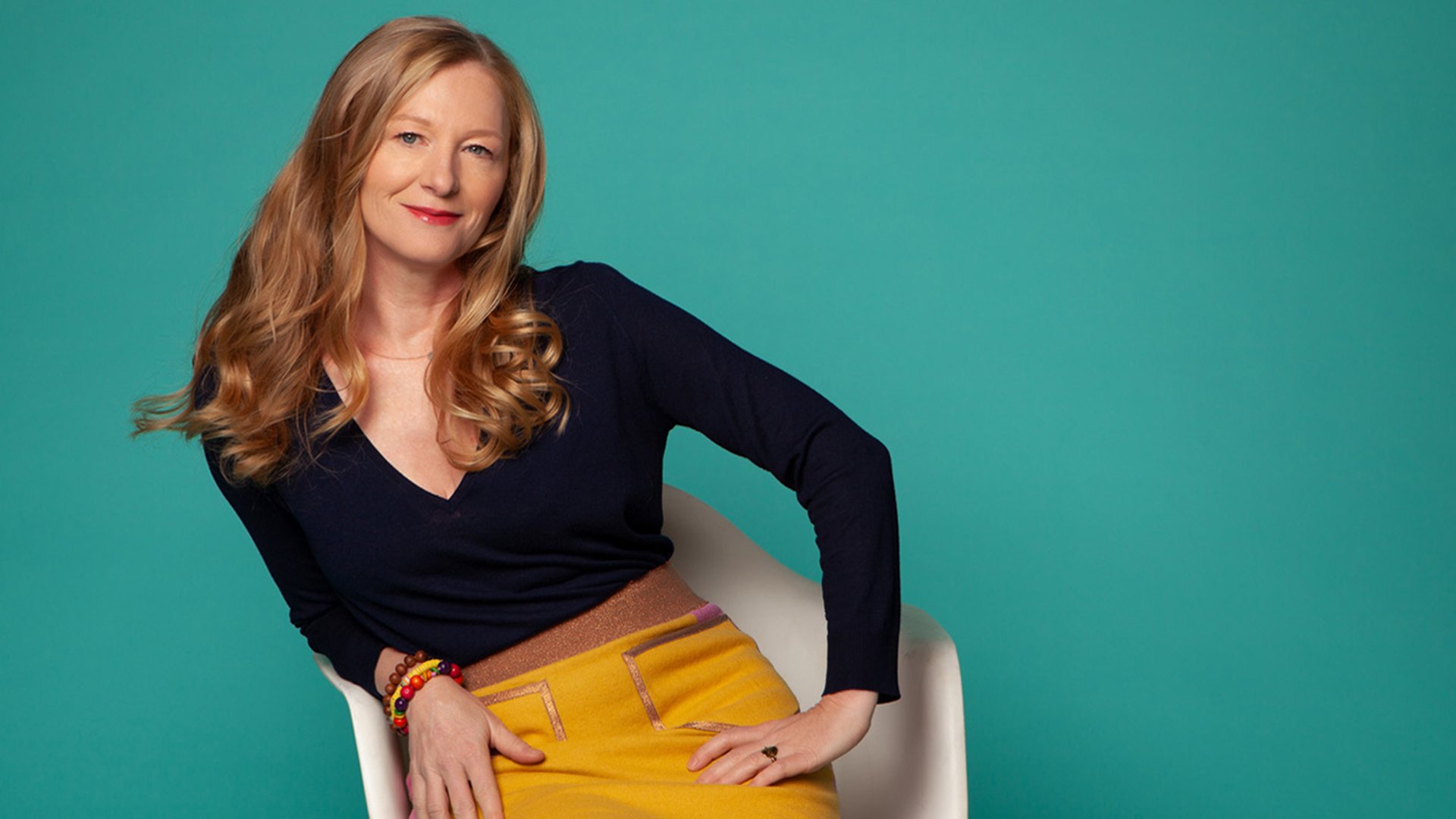 Lisa Wahlandt in gelbem Rock und schwarzem Oberteil auf einem Stuhl vor türkisfarbenem Hintergrund, mit locker gekreuzten Beinen und entspanntem Sitz.