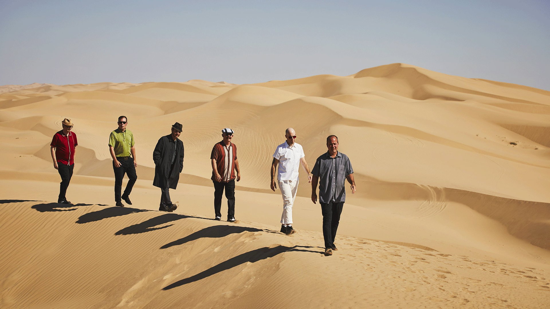 Bandfoto von The Slackers in einer Sandwüste. Sechs Bandmitglieder gehen in lockerer Formation über eine Dünenlandschaft und tragen unterschiedliche Freizeitkleidung sowie Hüte und Sonnenbrillen.