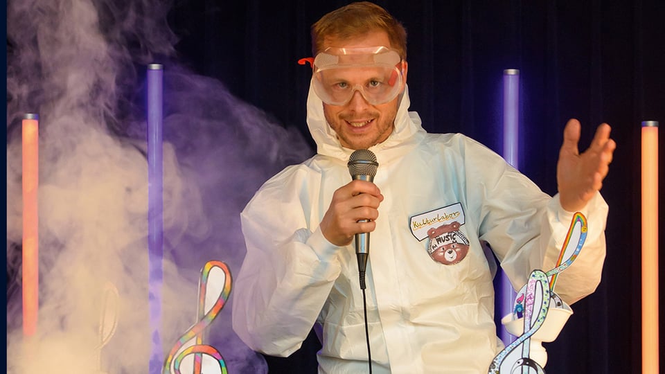 Bühnenfoto mit Person in weißem Schutzanzug und Schutzbrille, die ein Mikrofon hält, vor dunklem Hintergrund mit Nebel und leuchtenden farbigen Lichtstäben sowie bunten Notenschlüsselmotiven im Vordergrund.