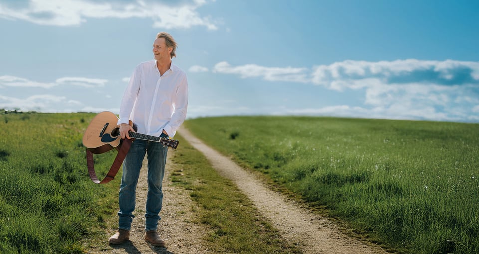 der Künstler mit weißem Hemd und Gitarre auf einer grünen Wiese