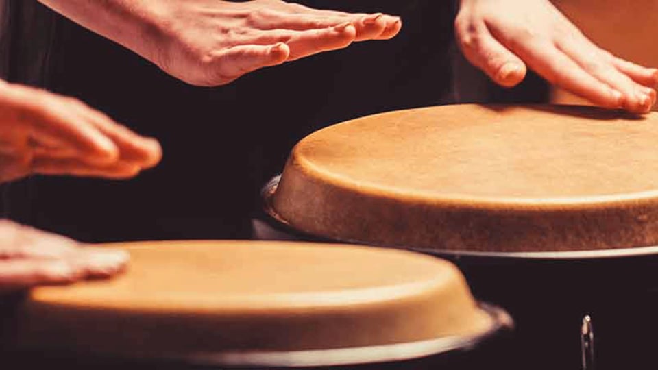 Nahaufnahme mehrerer Hände, die auf Conga Trommeln spielen. Die Aufnahme zeigt die Schlagbewegungen auf den naturfarbenen Trommelfellen vor dunklem Hintergrund.