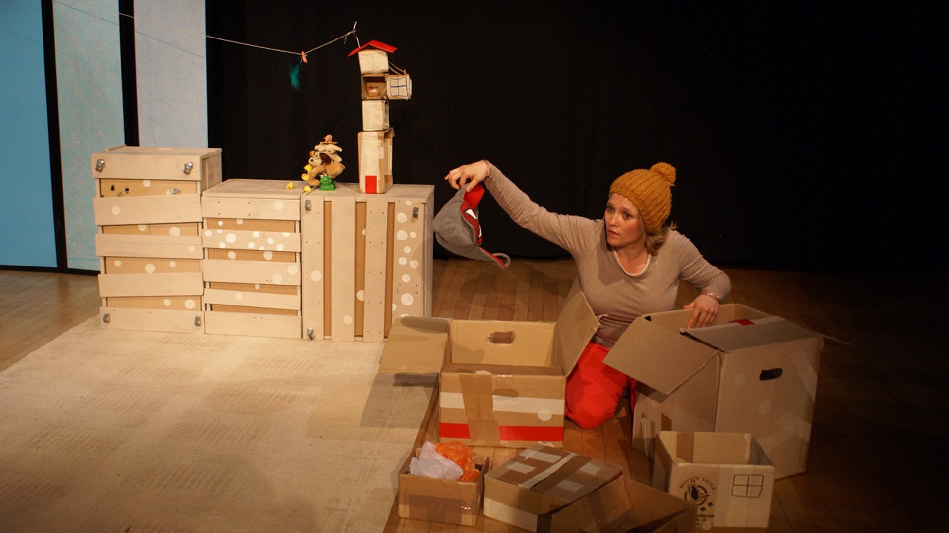 Foto für die Veranstaltung So weit oben, mit einer Frau, die auf dem Boden sitzt, umgeben von einigen Kartonschachteln.