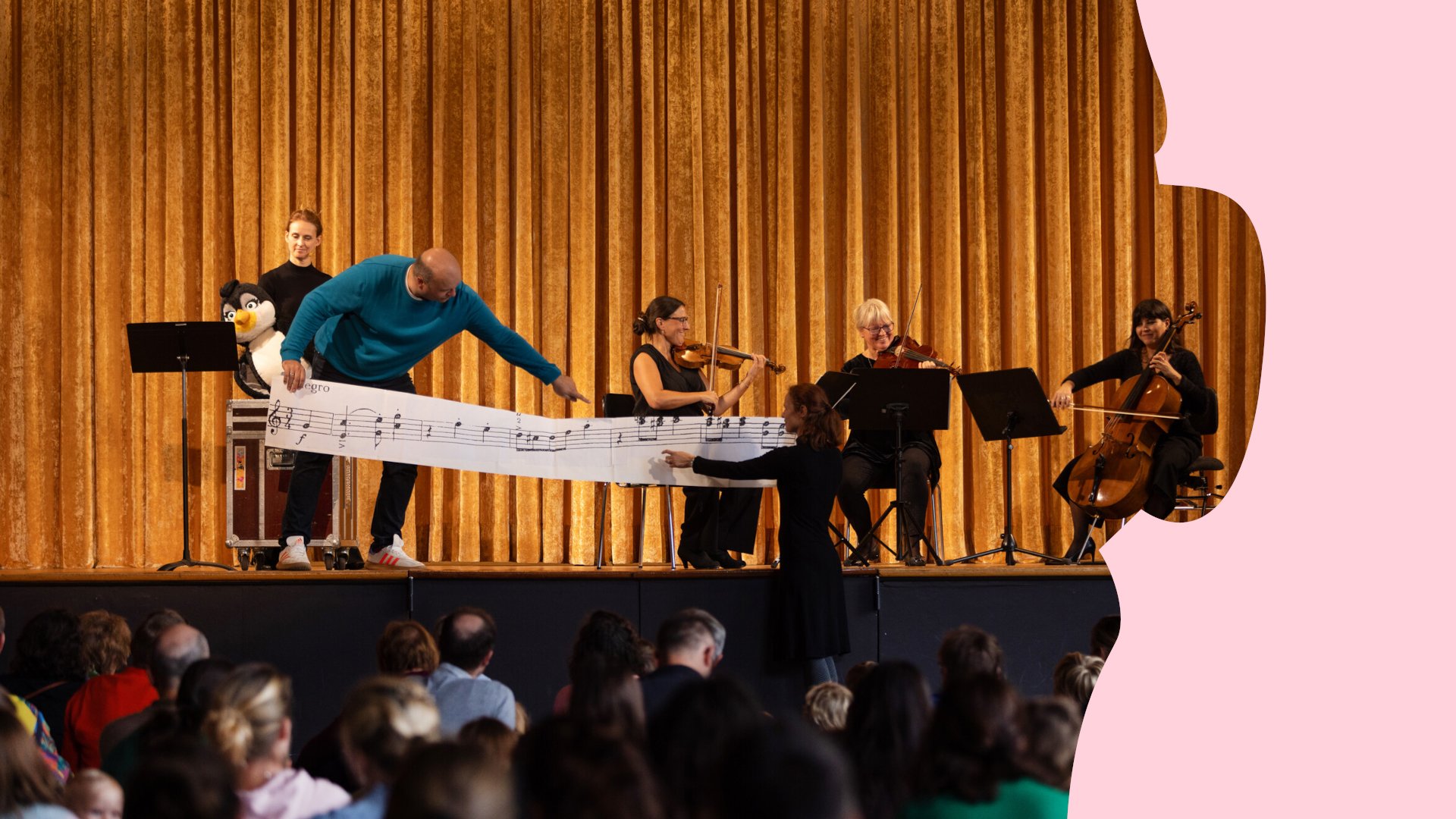 Musikalische Familienveranstaltung auf einer Bühne mit Streichquartett, Moderator und übergroßer Notenzeile vor goldenem Vorhang; Publikum im Vordergrund.