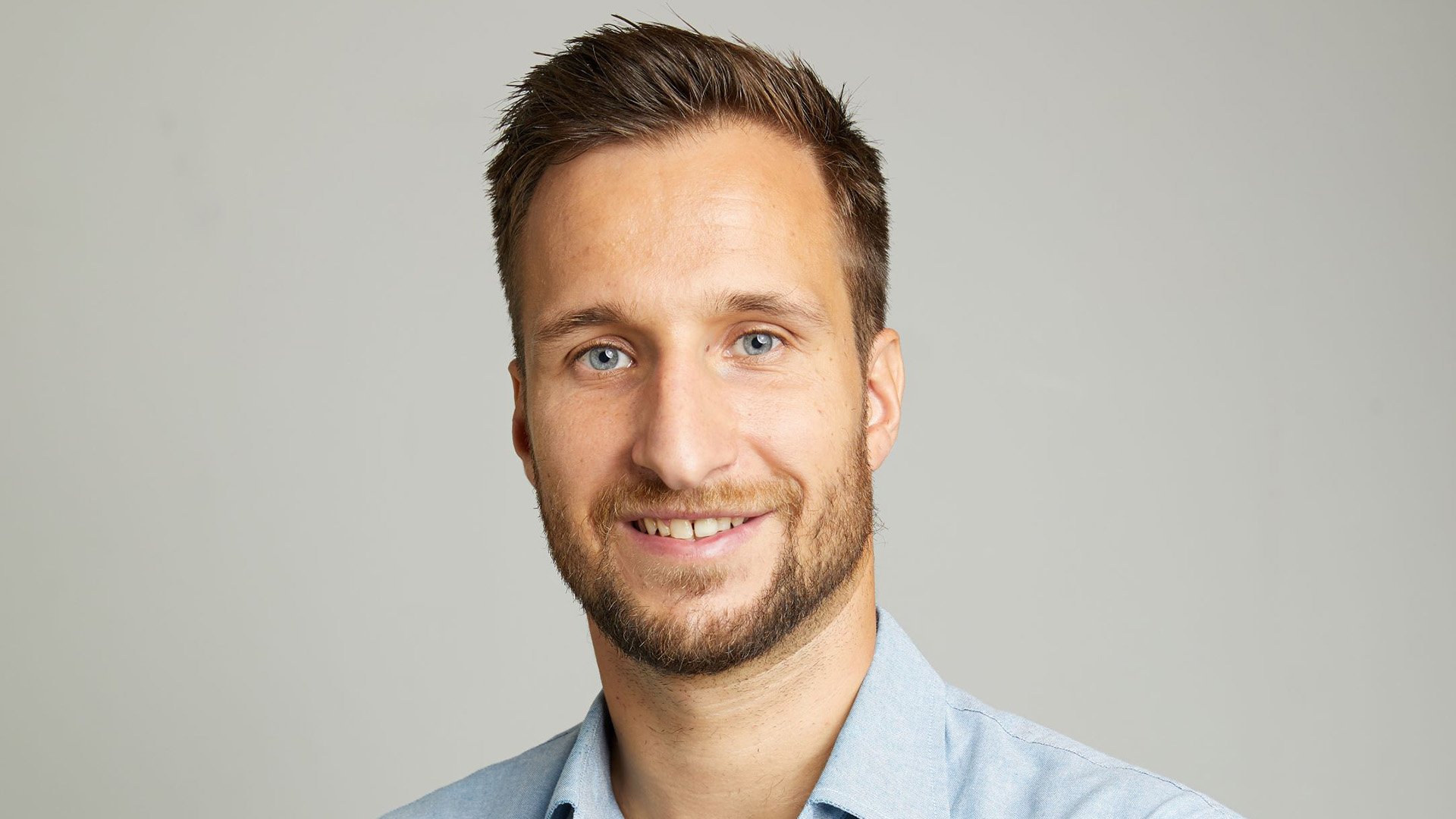Porträt einer Person mit kurzem Haar und Bart vor einem hellgrauen Hintergrund. Die Person trägt ein hellblaues Hemd und blickt direkt in die Kamera. Die Aufnahme ist als klassisches Kopf- und Schulterporträt gestaltet.