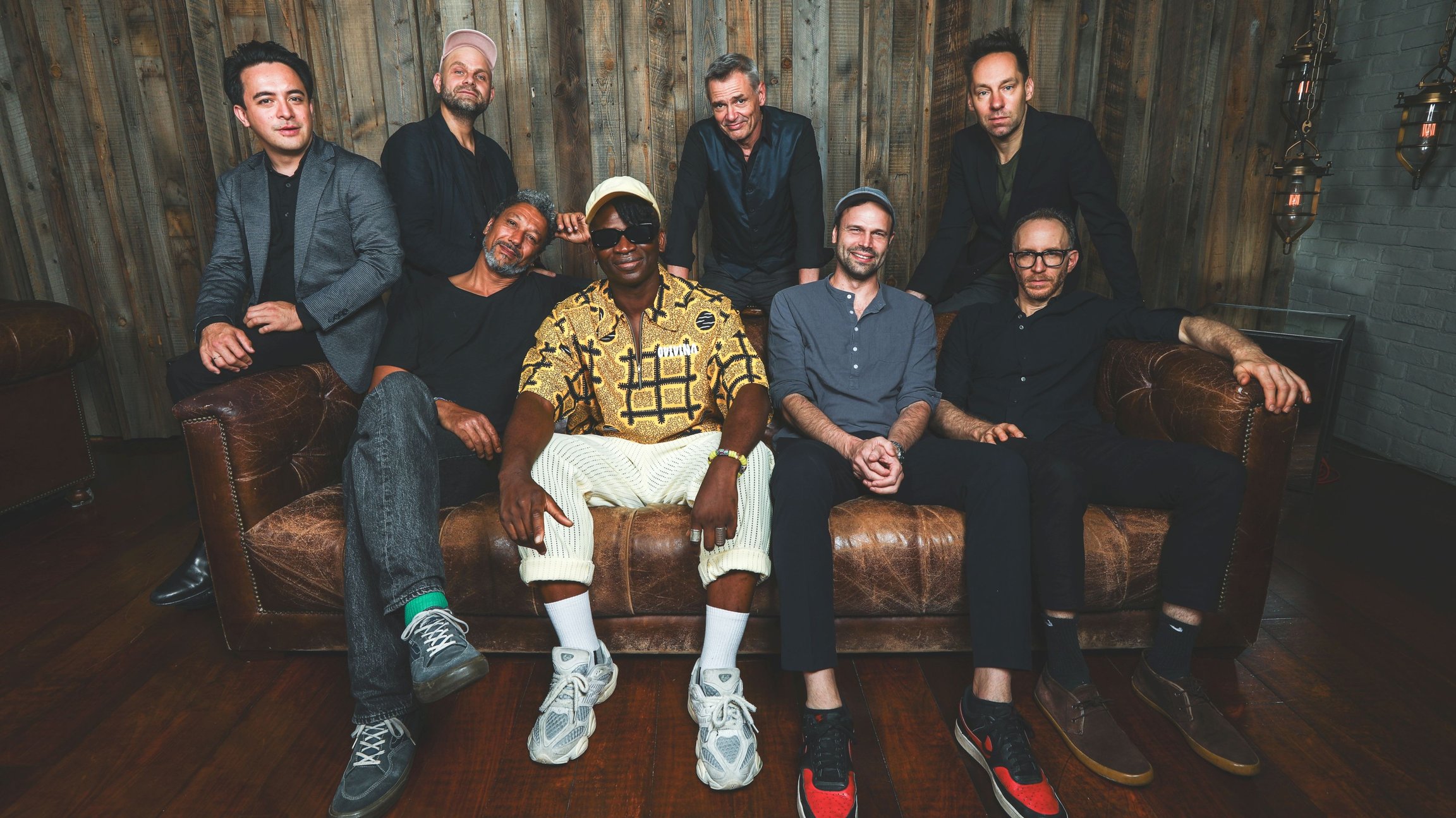 Acht Personen der Formation Jazzanova sitzen auf einem antiken Ledersofa vor einer Holzvertäfelung