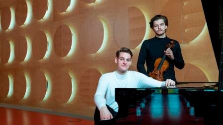 Zwei Musiker in einem modernen Konzertsaal neben einem Flügel. Eine Person sitzt am Klavier, die andere hält eine Violine; im Hintergrund ist eine geschwungene Wand mit kreisförmigen Elementen zu sehen.