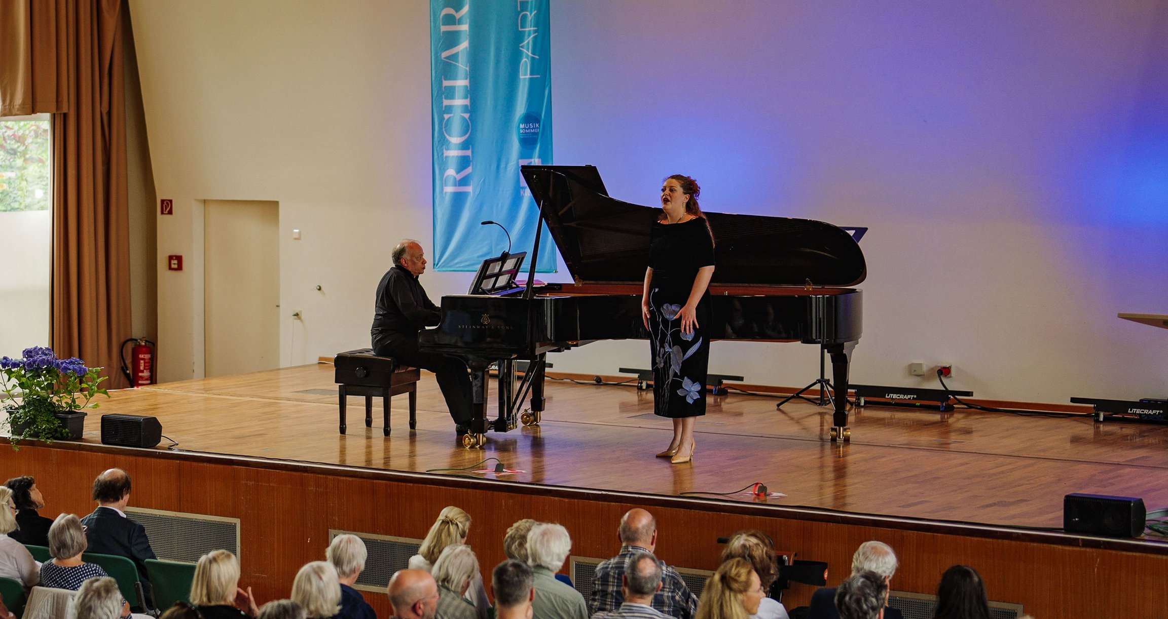 eine Sängerin steht auf einer Bühne undn wird von einem Klavier begleitet