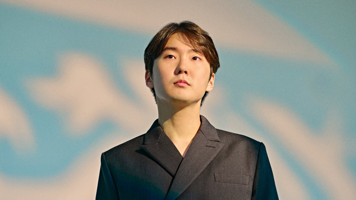 Der Künstler Seong Jin Cho in einem schwarzen Sakko vor hellblau-rosa verschwommenem Hintergrund