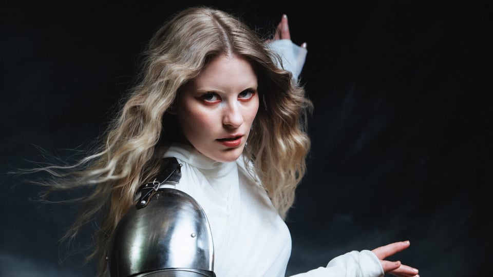 Person mit blondem, offenem Haar trägt ein weißes Oberteil und eine metallene Schulterrüstung und posiert vor dunklem Hintergrund.
