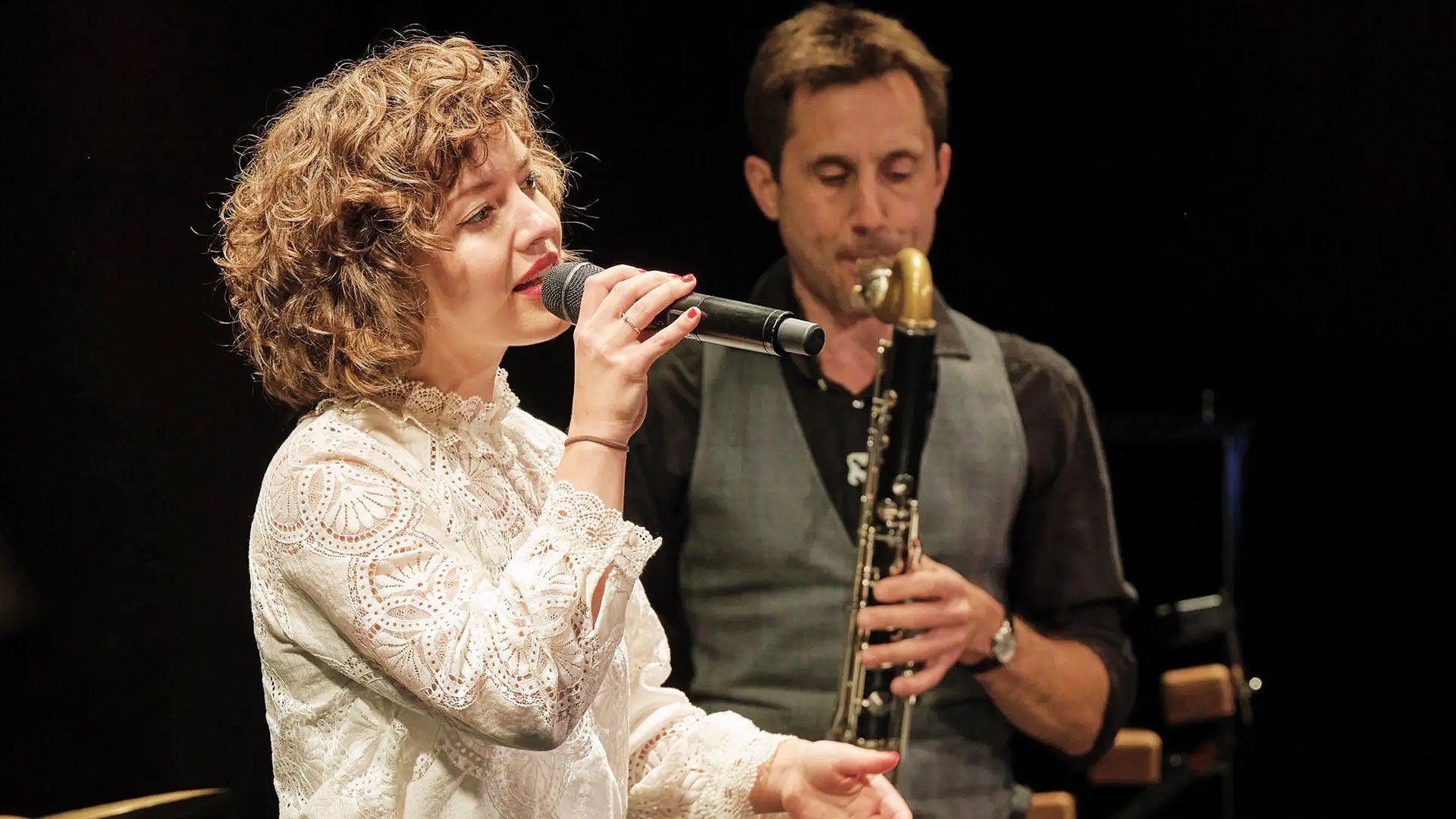 Bühnenfoto Sarah und das Nix mit Person im weißen Spitzenoberteil, die in ein Mikrofon singt, während im Hintergrund eine weitere Person Klarinette spielt, vor dunklem Hintergrund