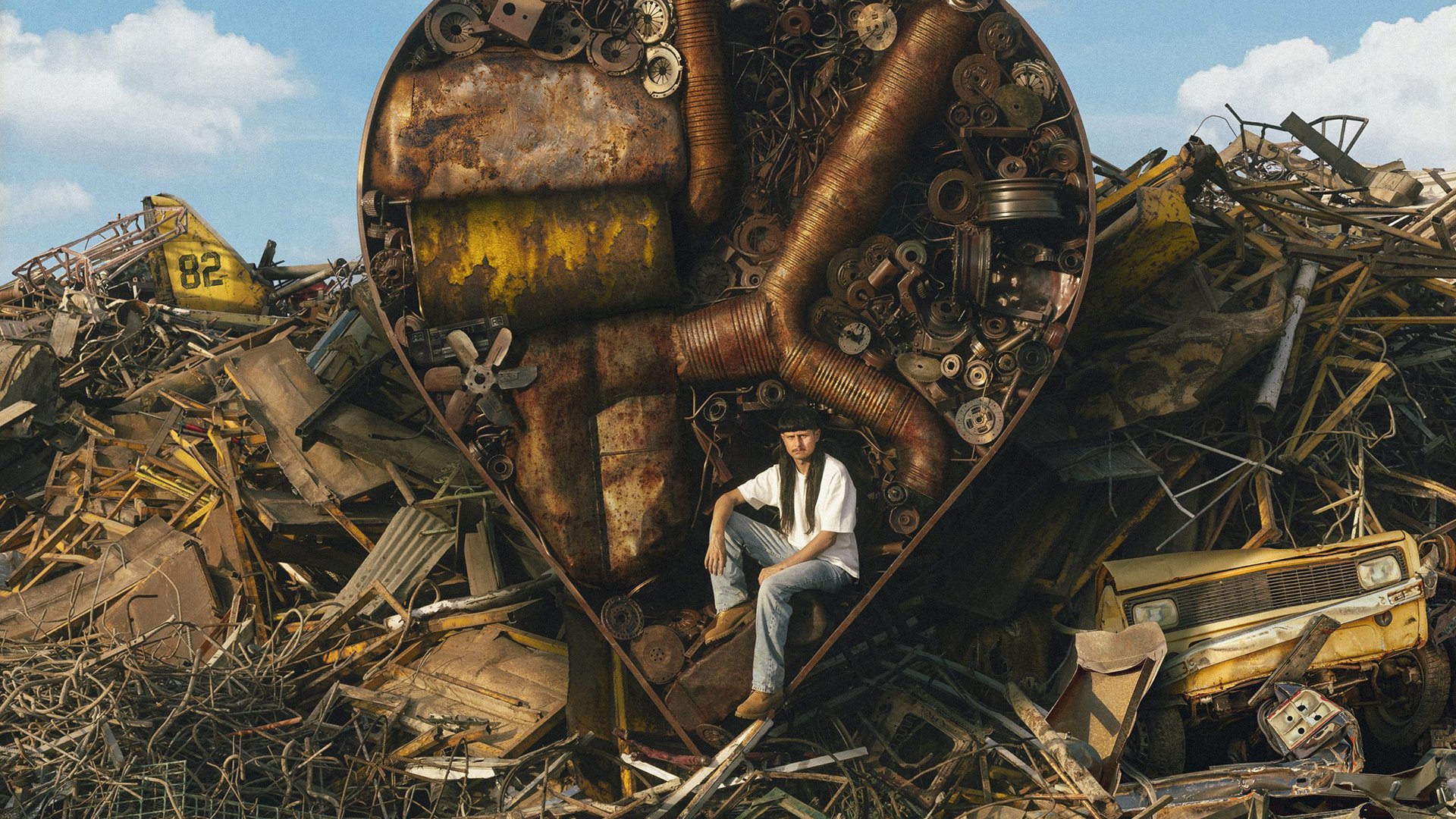 Veranstaltungsbild von Oliver Tree: Künstler sitzt inmitten eines Schrottplatzes vor einer großen, herzförmigen Skulptur aus rostigem Metall und mechanischen Bauteilen. Umgeben von Autoteilen, Zahnrädern und Industrieabfällen ist die Szene unter freiem Himmel inszeniert. Oliver Tree trägt ein helles T‑Shirt und Jeans und sitzt zentral im Bild. Die Bildgestaltung wirkt surreal und industriell und unterstreicht den alternativen, visuellen Stil des Musikprojekts.