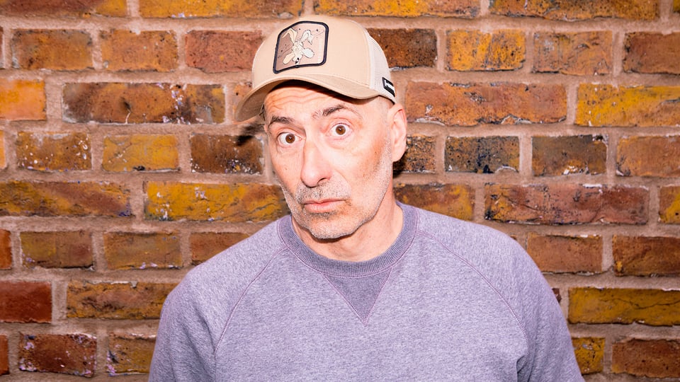 Eine Person trägt ein beigefarbenes Basecap mit aufgenähtem Patch sowie ein graues Sweatshirt und steht vor einer Wand aus rötlich‑braunen Backsteinen