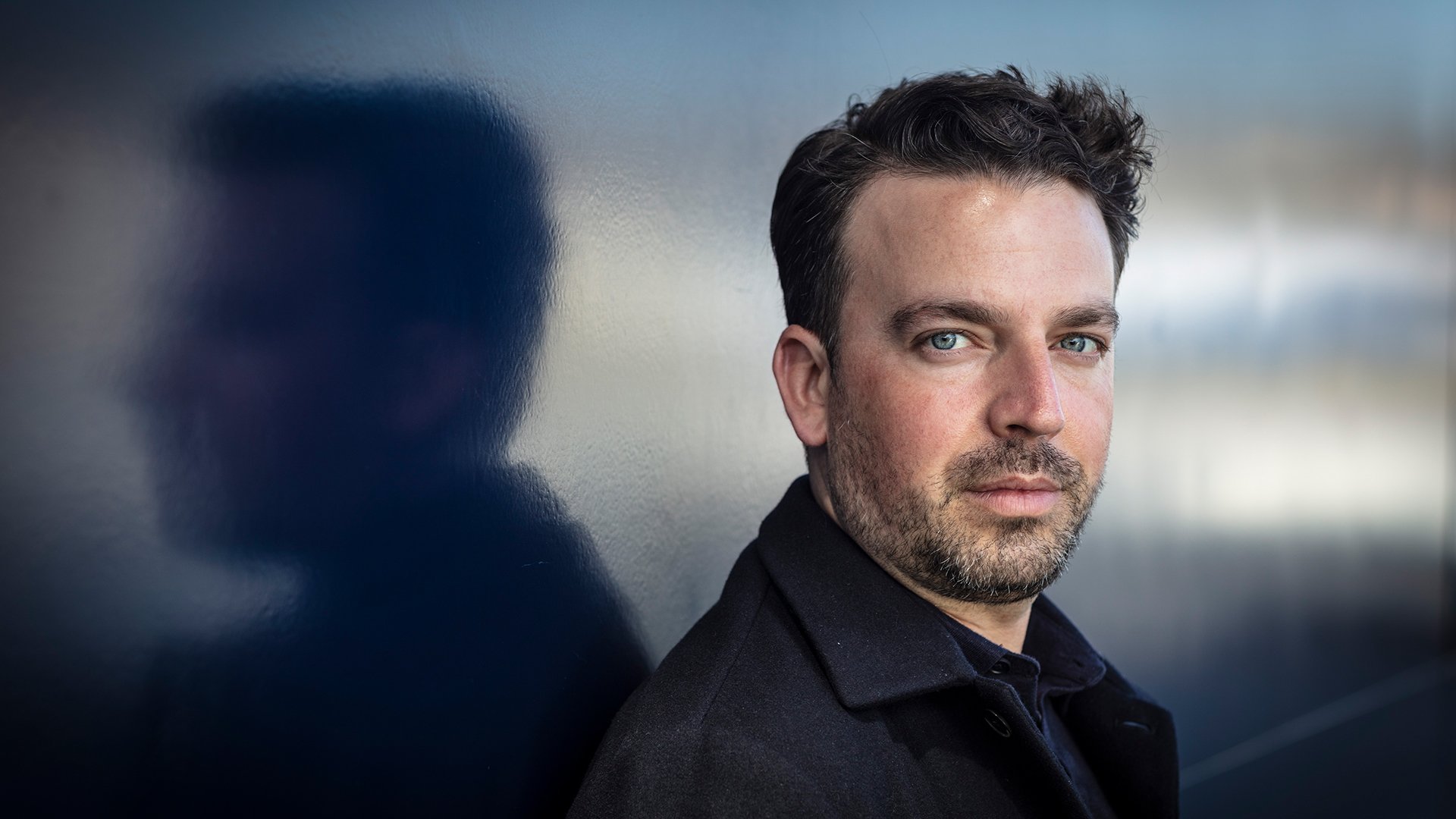 James Gaffigan in dunkler Kleidung vor einer Glaswand, seitlich aufgenommen mit Spiegelung und unscharfem Hintergrund in kühlen Farbtönen