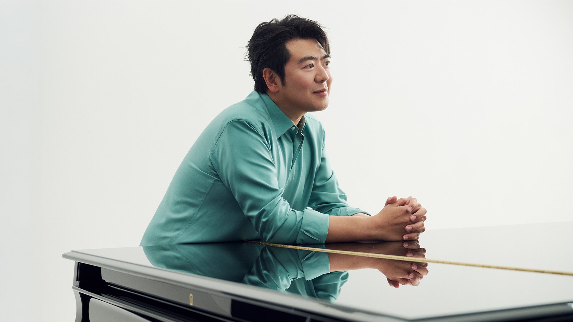 Porträtmotiv von Lang Lang: Der Pianist sitzt seitlich am Flügel und legt die gefalteten Hände auf den hochglänzenden Klavierdeckel. Er trägt ein türkisfarbenes Hemd. Der Hintergrund ist hell und neutral gehalten, ohne weitere Elemente. Ruhige, reduzierte Studioaufnahme; Promomotiv für einen klassischen Klavierabend mit Lang Lang in der Isarphilharmonie.