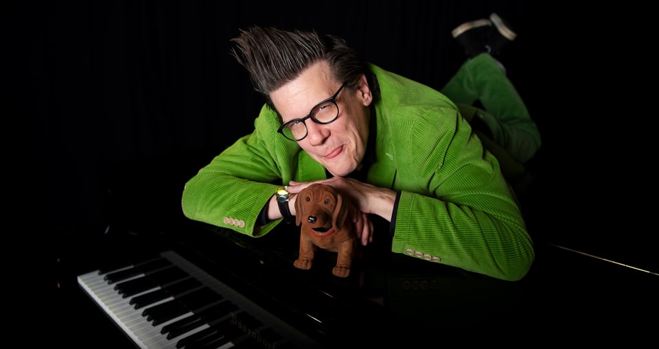 Ein Foto des Künstlers, der auf einem Klavier liegt, in Grün gekleidet, mit einem Plüschhund.