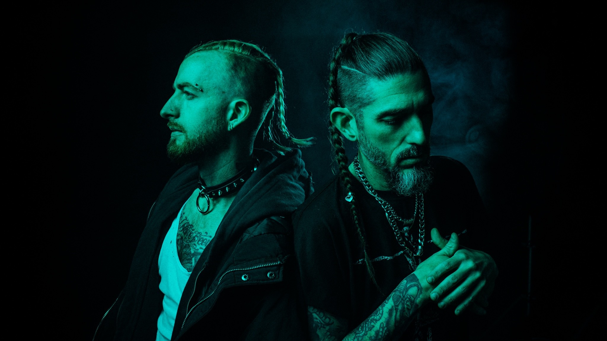 Die beiden Mitglieder von Missio mit Zöpfen, Halsketten und ärmelloser Kleidung stehen Rücken an Rücken vor dunklem Hintergrund, seitlich beleuchtet in grünem Licht