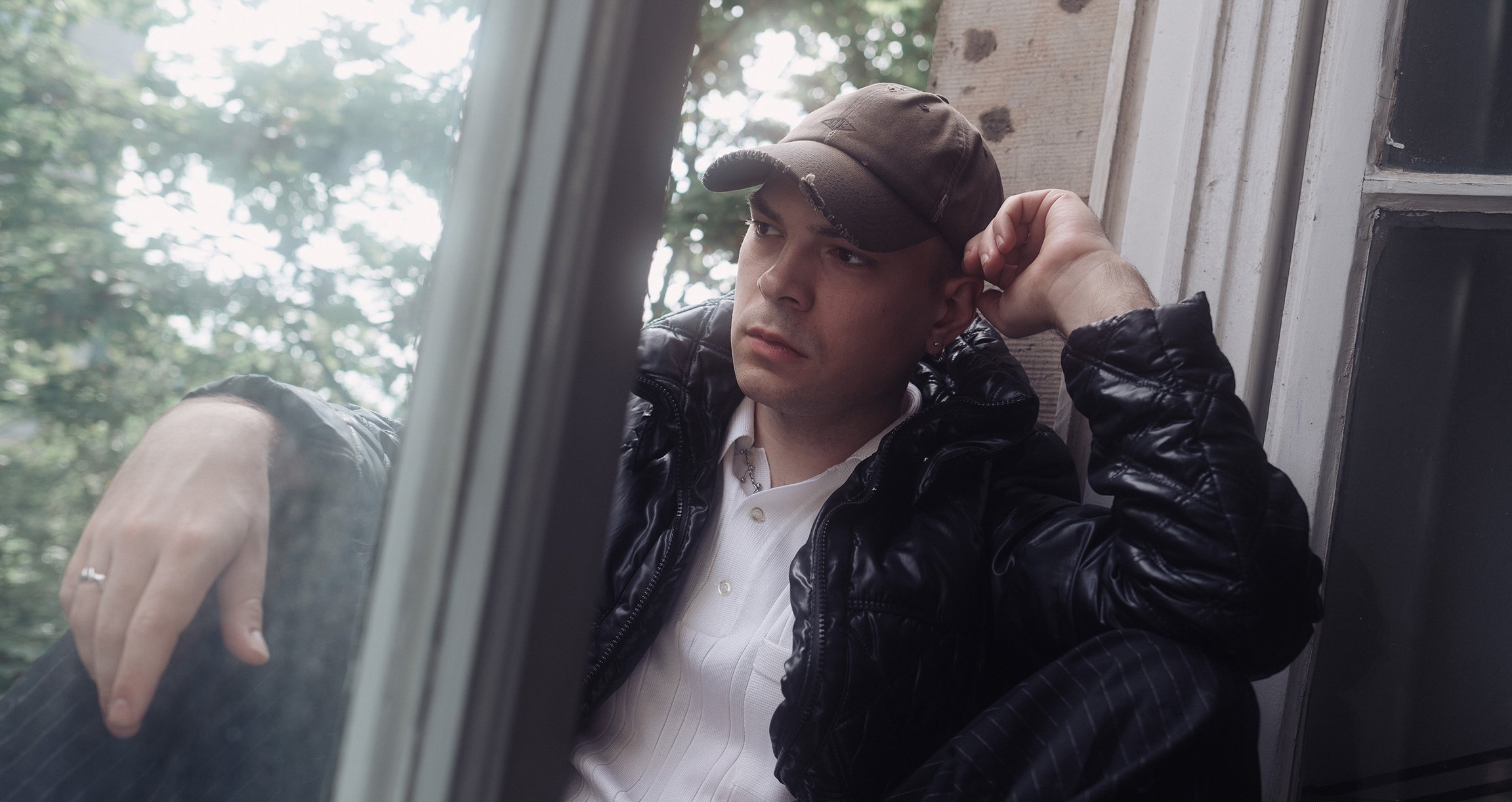 Der Künstler in einer schwarzen Jacke mit brauner Kappe sitzt an einem Fenster