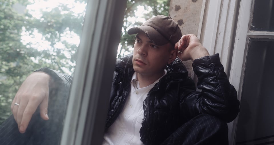 Der Künstler in einer schwarzen Jacke mit brauner Kappe sitzt an einem Fenster