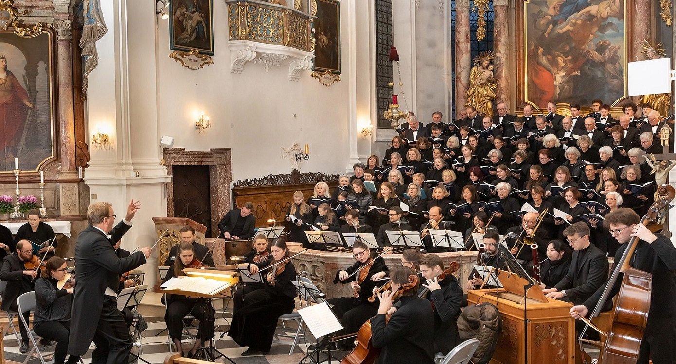 Orchester und großer Chor musizieren unter der Leitung einer dirigierenden Person in einer reich geschmückten Kirche.