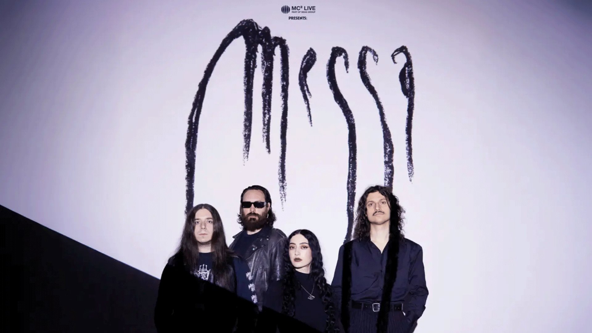 Bandfoto von Messa. Vier Bandmitglieder in dunkler Kleidung stehen vor hellem Hintergrund unter dem großflächigen Bandlogo Messa. Eine Person trägt eine Sonnenbrille und Lederjacke, während die Gruppe direkt in die Kamera blickt.
