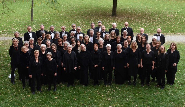 Der Chor in schwarzer Kleidung auf einer WIese, im Hintergrund Baumstämme und ein Weg