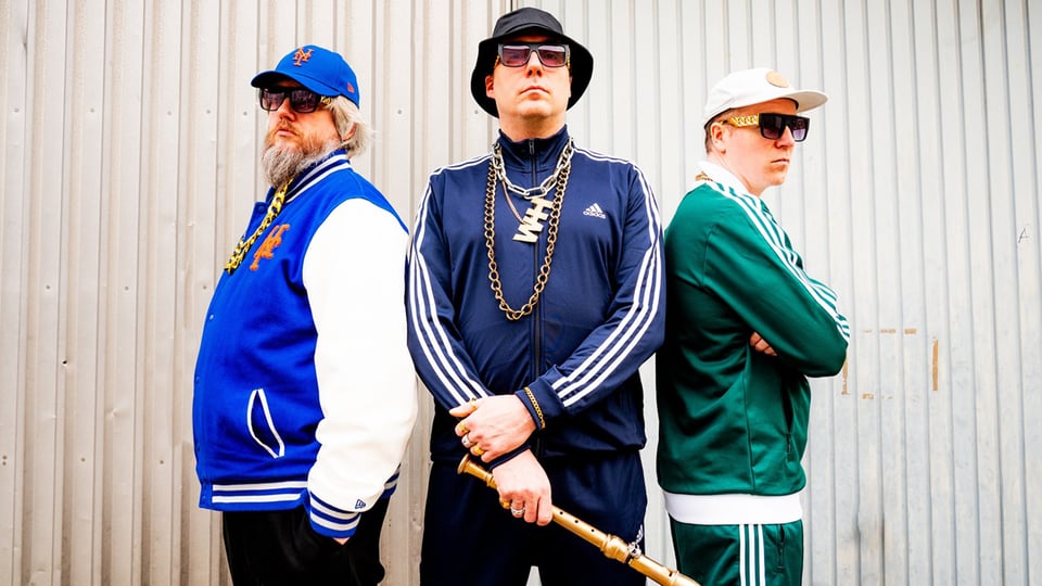 Drei Männer stehen mit Sonnenbrillen und Caps und Trainingsjacken im Kreis mit dem Rücken zueinander.