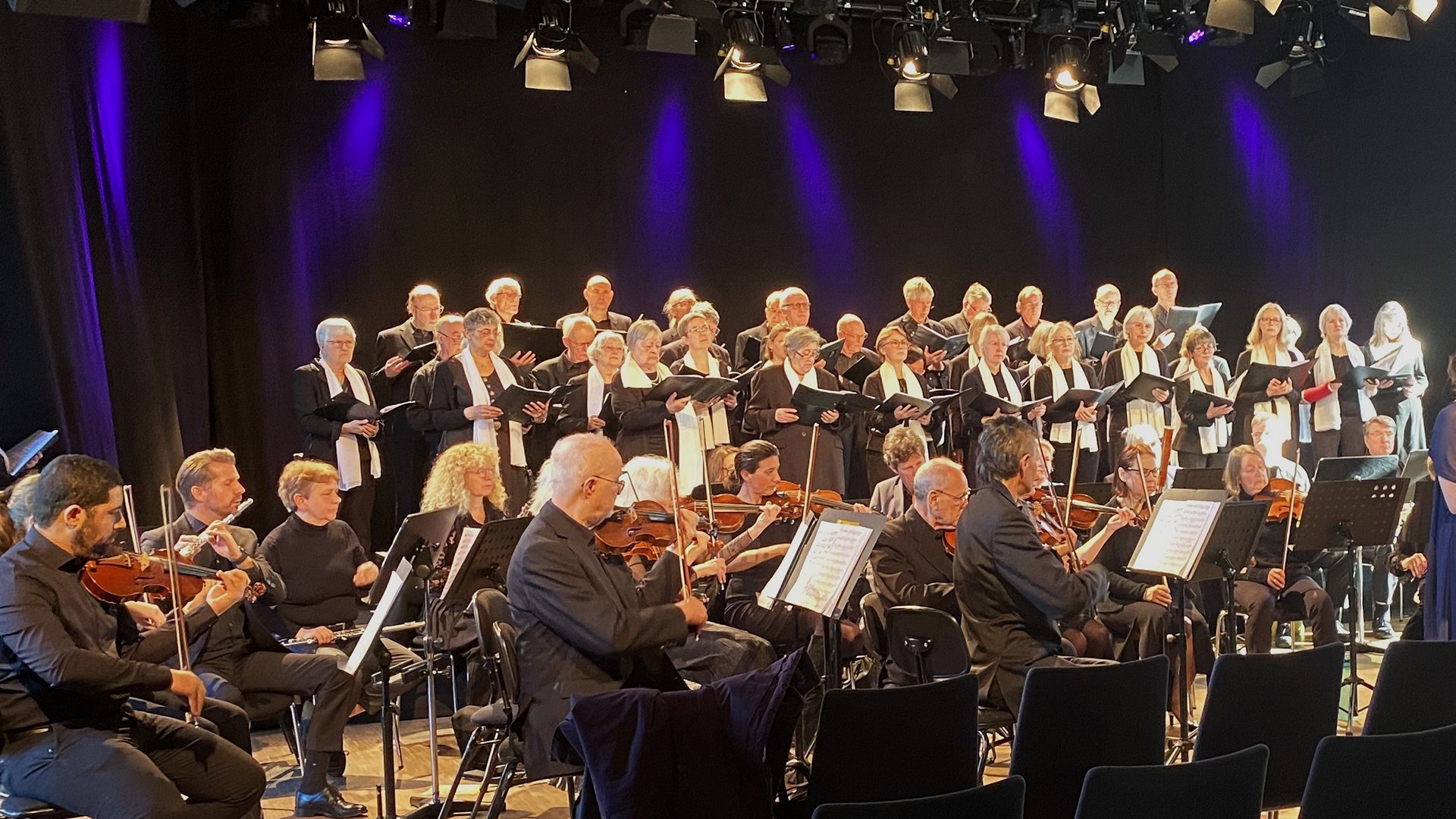 Veranstaltungsbild eines Chors mit Orchester bei einem klassischen Konzert in München: Auf einer Theaterbühne steht ein großer gemischter Chor mit Notenmappen in mehreren Reihen. Davor sitzt ein Orchester mit Streichinstrumenten wie Geigen und Bratschen sowie Holzbläsern an Notenpulten. Die Aufführung findet unter Bühnenbeleuchtung mit violetten und warmen Lichtakzenten statt. Das Ensemble ist in dunkler Konzertkleidung gekleidet und präsentiert ein musikalisches Programm im Rahmen einer Live‑Aufführung.