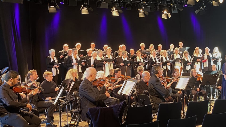 Veranstaltungsbild eines Chors mit Orchester bei einem klassischen Konzert in München: Auf einer Theaterbühne steht ein großer gemischter Chor mit Notenmappen in mehreren Reihen. Davor sitzt ein Orchester mit Streichinstrumenten wie Geigen und Bratschen sowie Holzbläsern an Notenpulten. Die Aufführung findet unter Bühnenbeleuchtung mit violetten und warmen Lichtakzenten statt. Das Ensemble ist in dunkler Konzertkleidung gekleidet und präsentiert ein musikalisches Programm im Rahmen einer Live‑Aufführung.