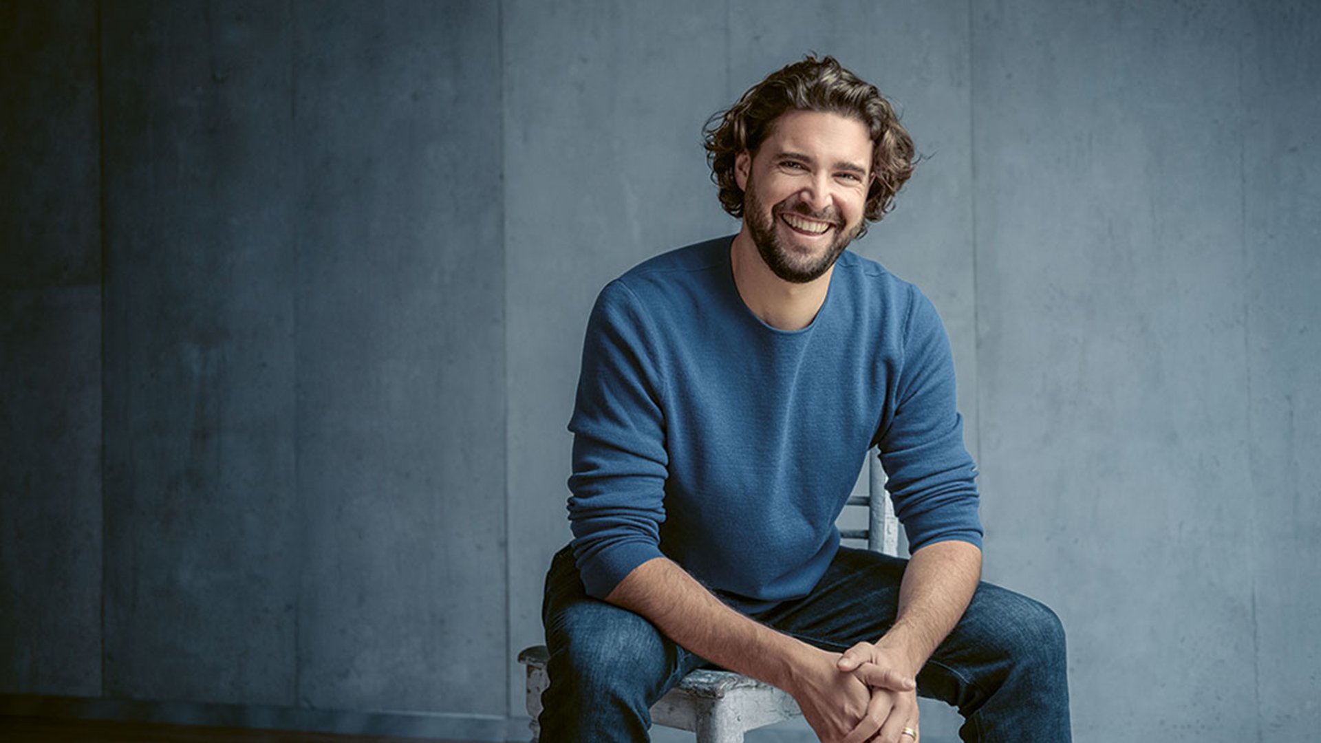 Christian Reif in blauem Pullover sitzt lachend auf einem Hocker vor blau-grauem Hintergrund